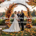 Bride and groom holding hands under a vibrant floral arch during a fall outdoor wedding ceremony, surrounded by autumn foliage and guests' chairs decorated with flowers.